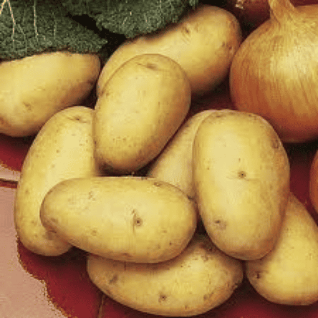 A close-up pile of oval yellow potatoes with a red surface, partially surrounded by a yellow onion and dark green leafy greens.